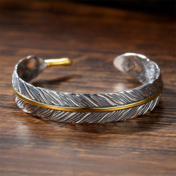 Open cuff feather bracelet with silver body and gold rachis accent, interior engraved scrollwork on polished dark wood background