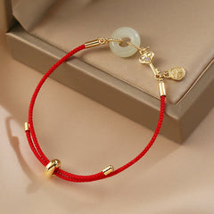 Adjustable red silk cord bracelet with celadon jade pi disc pendant, gold ruyi scepter charm with crystal and fortune medallion on beige jewelry box
