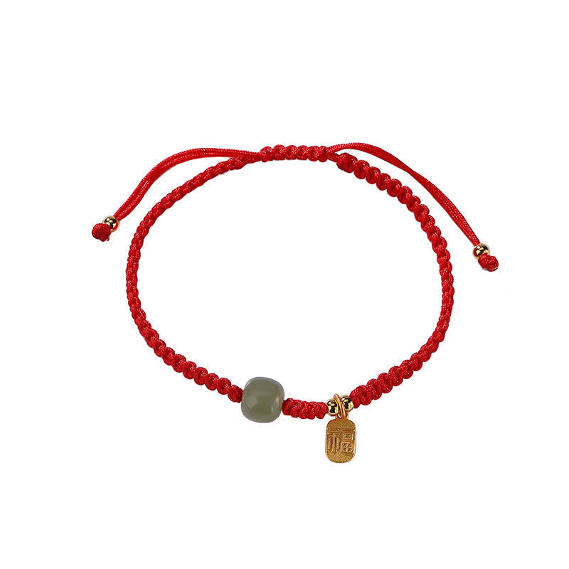 Black cord adjustable bracelet with Hetian jade green bead rectangular gold fu fortune charm and gold-toned spacer beads on taupe textured surface with warm wood prop accent