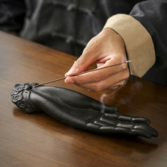 Buddha Hand Incense Burner – hand inserting incense stick into matte black ceramic holder, cloud-pattern base, serene