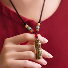 Vintage brass cylinder pendant necklace with green jade and amber beads held by hand, Tibetan Buddhist amulet on brown cord, red background