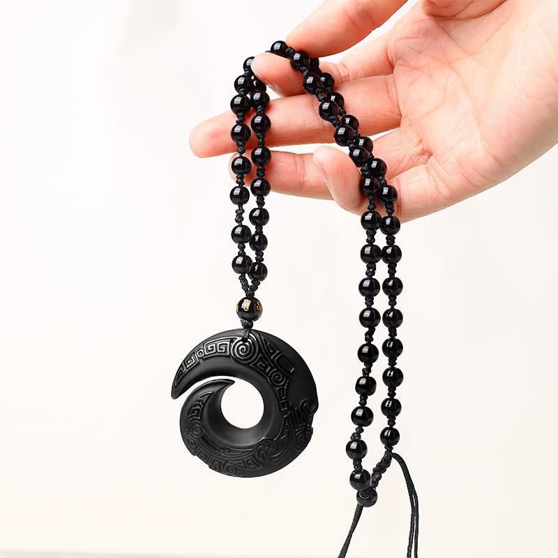 Black Obsidian Ice Obsidian Pendant Necklace – Hand-held black obsidian pendant necklace showing full beaded cord with gold inlaid character bead, circular spiral medallion with carved geometric patterns, white studio backdrop, unisex spiritual protection jewelry