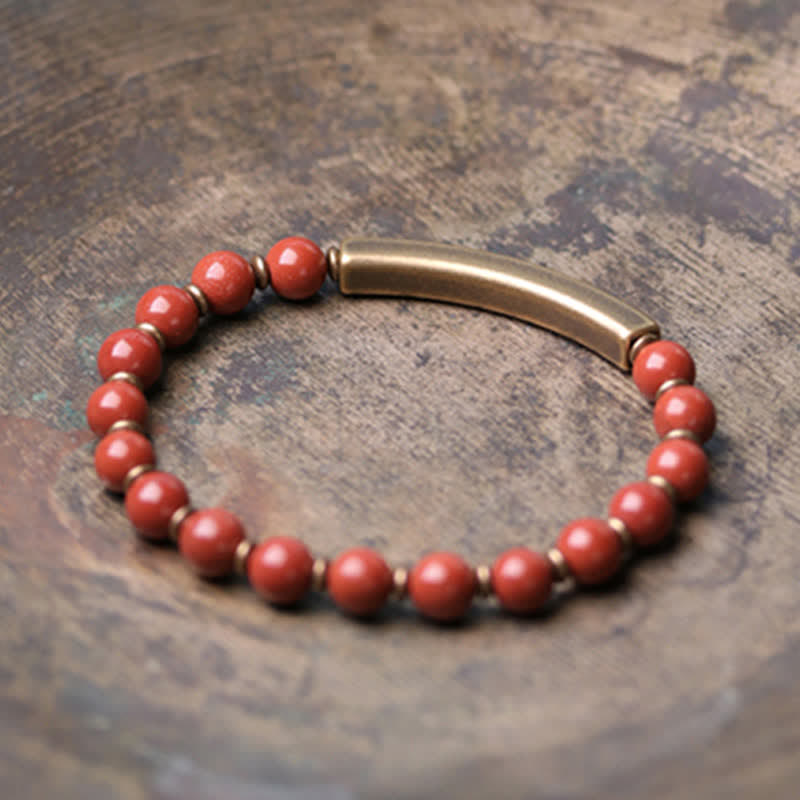 Red jasper bracelet on aged copper patina surface, burnt orange stone beads with smooth brass curved bar and gold-toned spacers, artisan jewelry photography