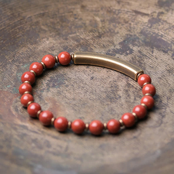 Red jasper bracelet on aged copper patina surface, burnt orange stone beads with smooth brass curved bar and gold-toned spacers, artisan jewelry photography