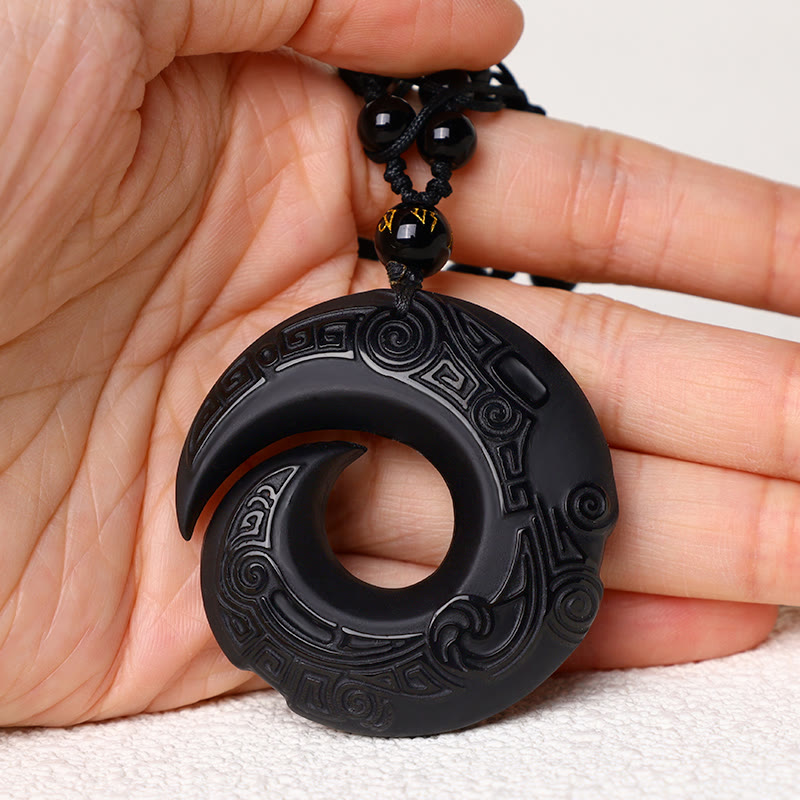 Black Obsidian Ice Obsidian Pendant Necklace – Black obsidian carved spiral pendant necklace held in hand for scale reference, showing intricate triskelion relief engraving and bi disc form, gold character bead, white fabric background, handmade natural stone amulet
