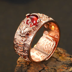 PiXiu Garnet Heart Sutra Ring – Silver foo dog ring with red gemstone and ornate relief band on dark rough rock surface, elevated frontal three-quarter view