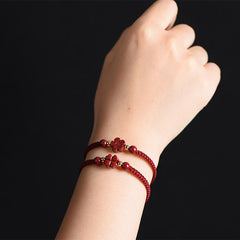 Two stacked burgundy braided cord bracelets with carved red four-leaf clover charms, gold accent beads and matching red stone beads worn on wrist, black background