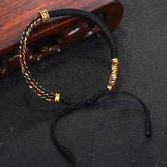 Om Mani Padme Hum Red String Bracelet – Black and multicolor woven bracelet with gold carved spacers on black matte background with carved wood prop