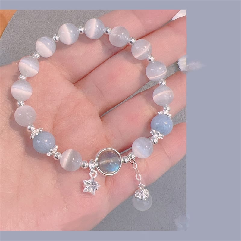 Cat's Eye Moonstone Butterfly Bracelet – White cat's eye beads with iridescent center and star crystal dangle charm on open palm, gray fabric blue frame border