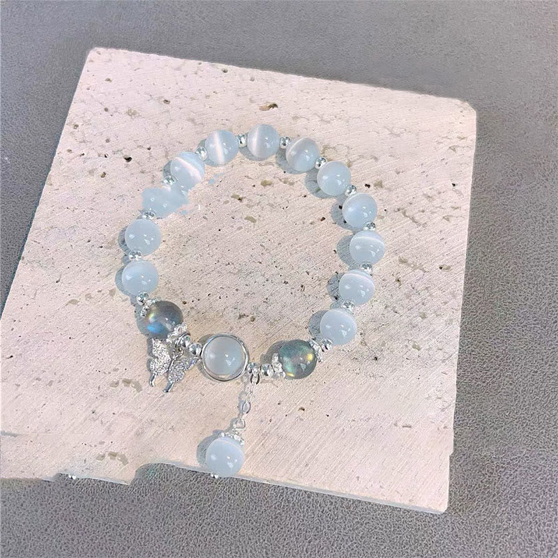 Cat's Eye Moonstone Butterfly Bracelet – Pale blue cat's eye beads with silver butterfly charm and bead drop on beige stone slab against gray fabric, top-down