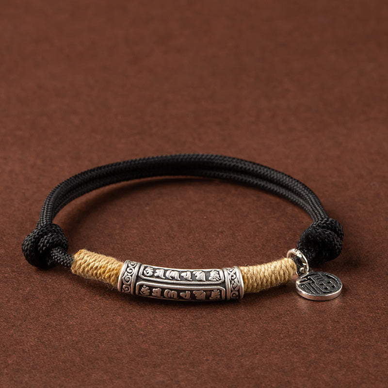 Silver mantra tube bracelet with beige and black braided cord worn on wrist, antique silver cylindrical charm with engraved details against burgundy fabric