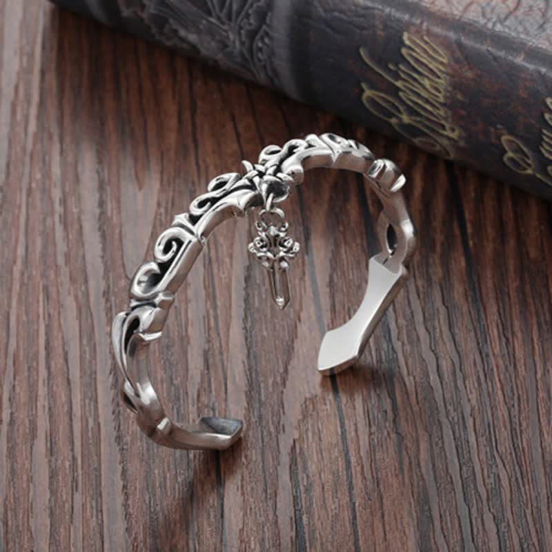Oxidized silver Viking cuff bracelet with ornate scrollwork fleur-de-lis motif and sword charm pendant displayed on dark wood with antique book background