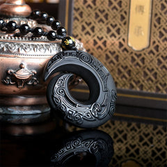 Black Obsidian Ice Obsidian Pendant Necklace – Three-quarter view of black obsidian spiral pendant with crescent moon cutout and archaic Chinese carved motifs, gold mantra bead accent, on black mirror surface with antique bronze vessel and traditional lattice panel, protective talisman jewelry