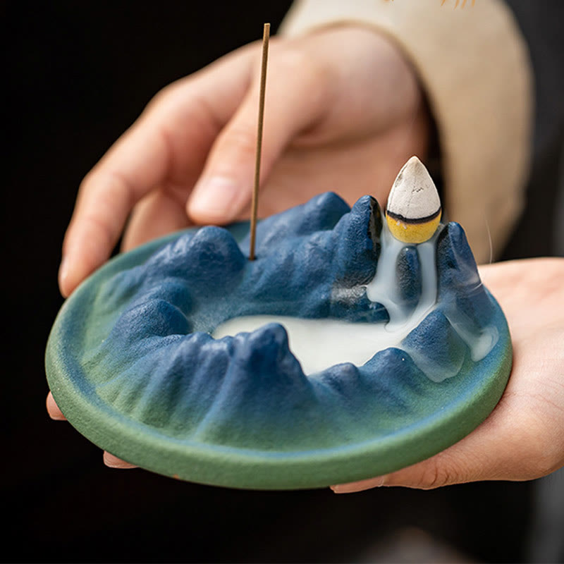 Ceramic Backflow Incense Burner – Hands holding teal mountain with white smoke waterfall against black, meditation