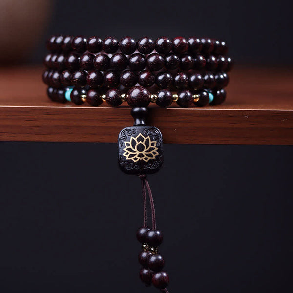 108 bead mala bracelet with polished red sandalwood beads, turquoise accent stones, gold-toned spacer beads and carved wood guru bead with gold lotus flower inlay on dark wood surface