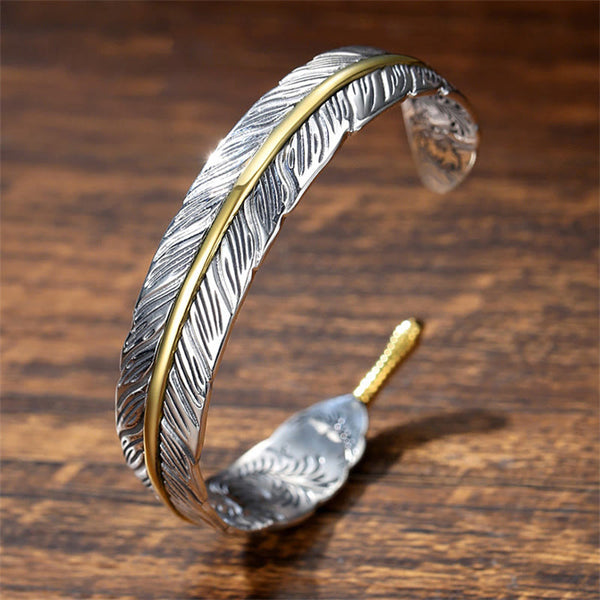 Feather-shaped silver cuff bracelet with gold central spine accent, oxidized antique finish and detailed barb etching on warm wood surface