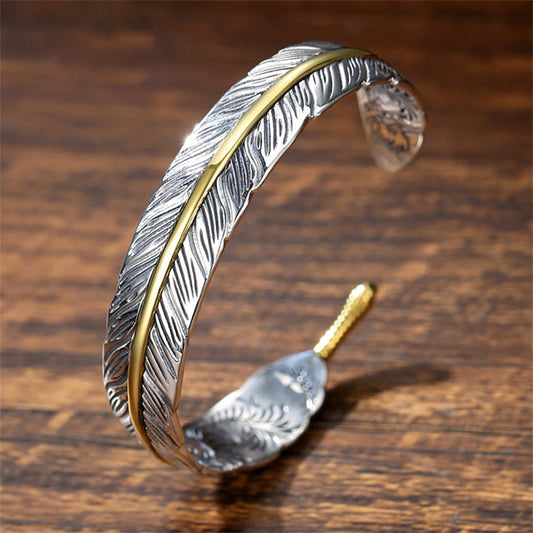 Feather-shaped silver cuff bracelet with gold central spine accent, oxidized antique finish and detailed barb etching on warm wood surface