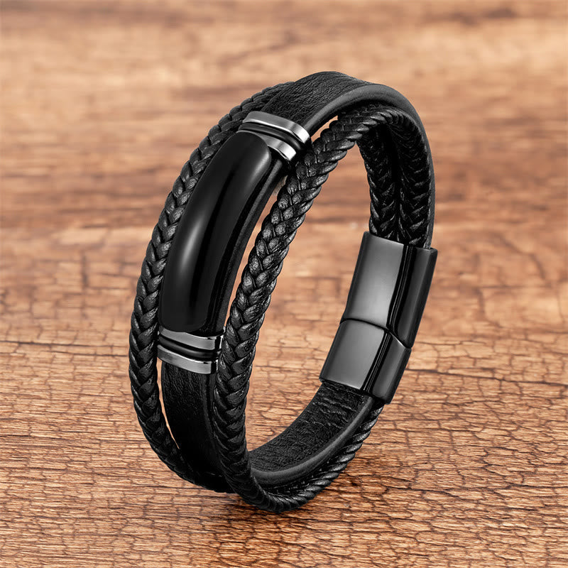 Triple-strand black leather bracelet with smooth center band and braided edges, polished gunmetal magnetic clasp, all-black design on wood surface.