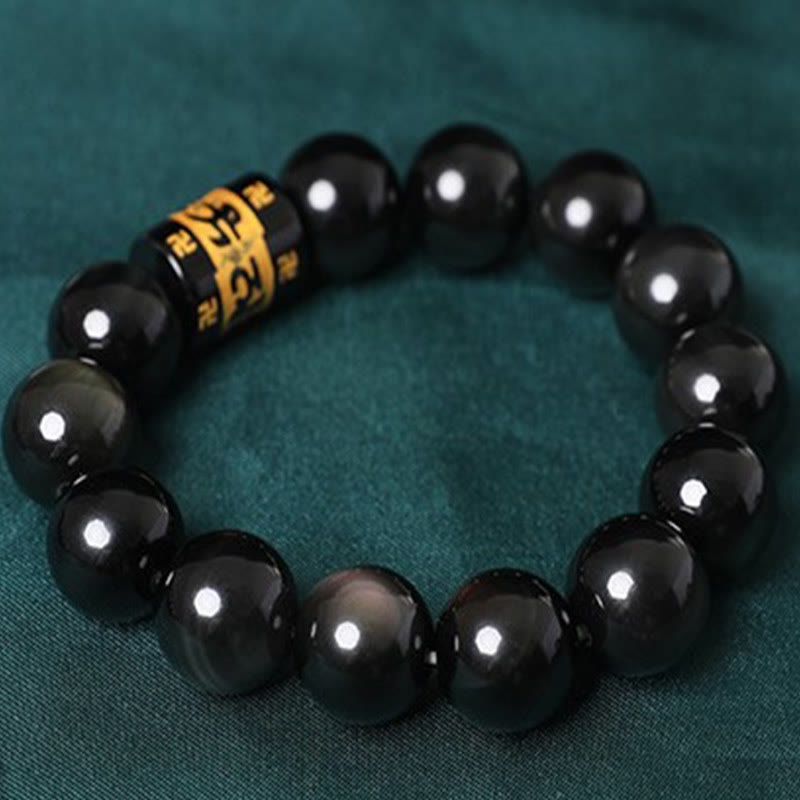 Rainbow Obsidian Bracelet – Top-down shot of polished black beads with gold mantra cylinder on green fabric, meditation beads