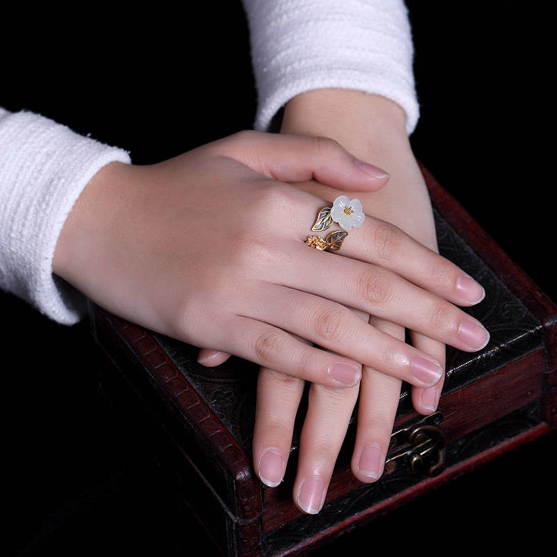 White Jade Plum Blossom Silver Ring - Ring on hand resting on ornate dark wooden box, showing white jade flower with gold stamen and silver leaf elements