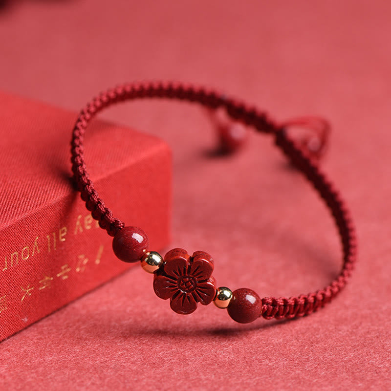 Deep red braided cord bracelet with carved cinnabar flower charm, gold accent beads displayed on red fabric with romantic gift box prop