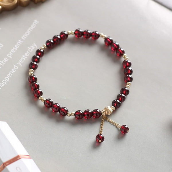 Natural garnet bracelet with 14K gold spacers and decorative dangle charms on light gray surface, gift box with rose gold ribbon visible, inspirational text background