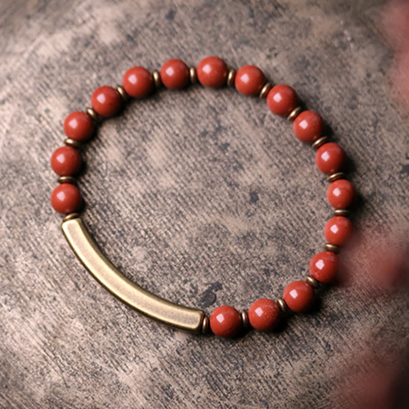 Red jasper bracelet on dark concrete background, coral-orange beads with antique brass curved bar and disc spacer beads, boho minimalist product shot