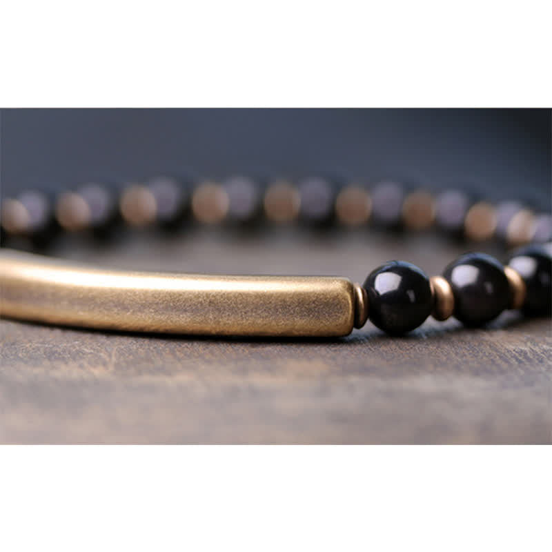 Black obsidian bracelet close-up with shallow depth of field, glossy black beads and antique brass curved bar on textured stone surface, minimalist men jewelry detail