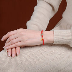 Red cord bracelet worn on model wrist against rust brown background showing Hetian jade green bead gold fortune fu charm pendant with cream knit sweater lifestyle styling