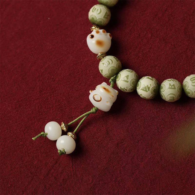 Bodhi Seed Bracelet – Angled shot of green beads, white cat charms and cord tassel on deep red fabric, charm bracelet