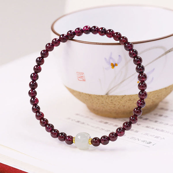 Natural garnet bracelet with jade focal bead resting on open book with Chinese characters and hand-painted ceramic teacup, traditional cultural styling