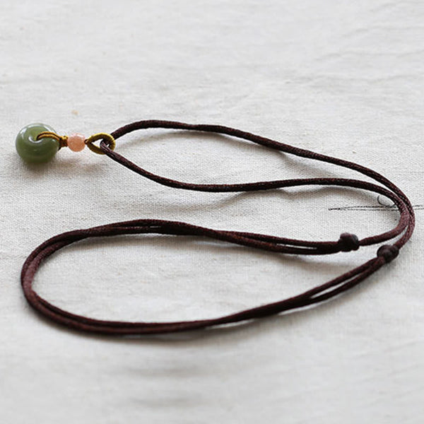 Jade Peace Buckle Lotus Necklace – Flat lay of jade donut on brown cord with adjustable knots on cream linen, jade necklace