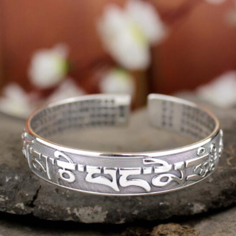 Tibetan Om Mani Padme Hum silver prayer bangle with raised Tibetan mantra, auspicious symbol motifs, micro-engraved interior, on slate with bokeh flowers.