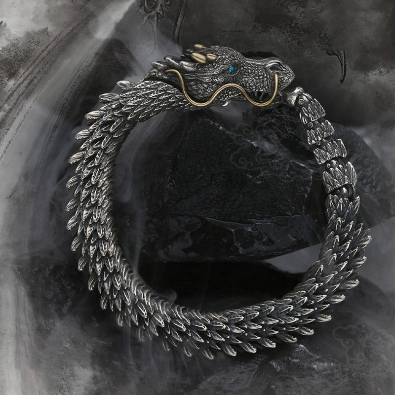 Coiled dragon ouroboros bracelet featuring articulated scale segments gold horn accents blue gemstone eyes and antiqued silver finish on dark background