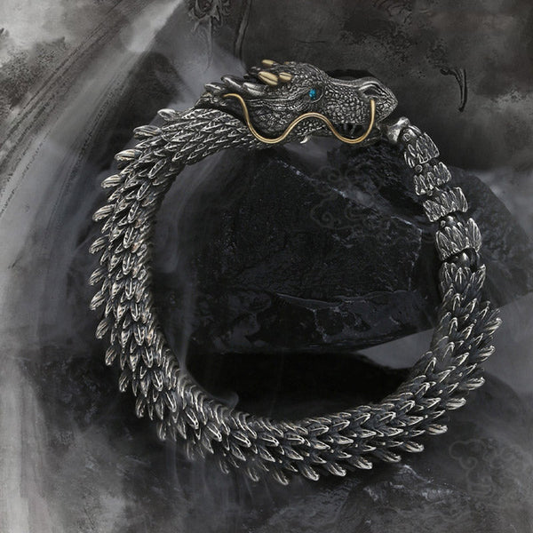 Coiled dragon ouroboros bracelet featuring articulated scale segments gold horn accents blue gemstone eyes and antiqued silver finish on dark background