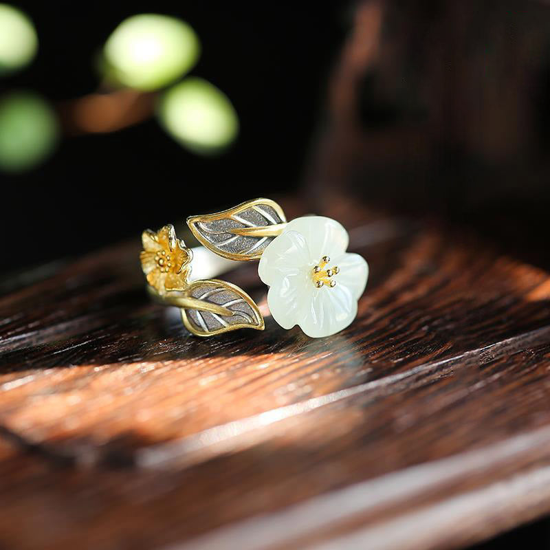 White Jade Plum Blossom Silver Ring - Ring on dark polished wood with pale celadon-white jade flower, gold stamen beads, gold flower bud, and oxidized silver leaves