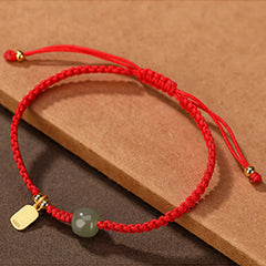 Adjustable red string bracelet with Hetian jade bead barrel-shaped green stone gold fu fortune charm and sliding knot closure on kraft paper and dark gray textured display
