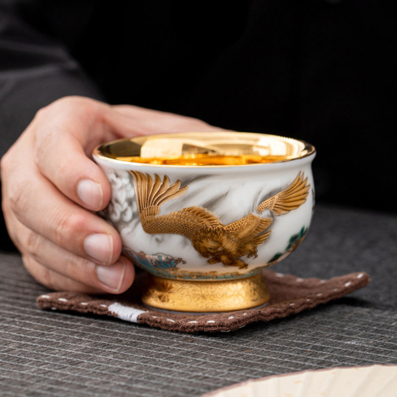Golden Eagle Dragon Gilt Ceramic Teacup – golden eagle relief with mountain landscape and gold pedestal base, held in hand