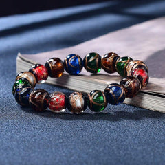 Colorful Liuli Glass Bead Luck Bracelet – Translucent rainbow glass beads with pale blue focal bead on gray-blue velvet cushion with gold decorative boxes, overhead