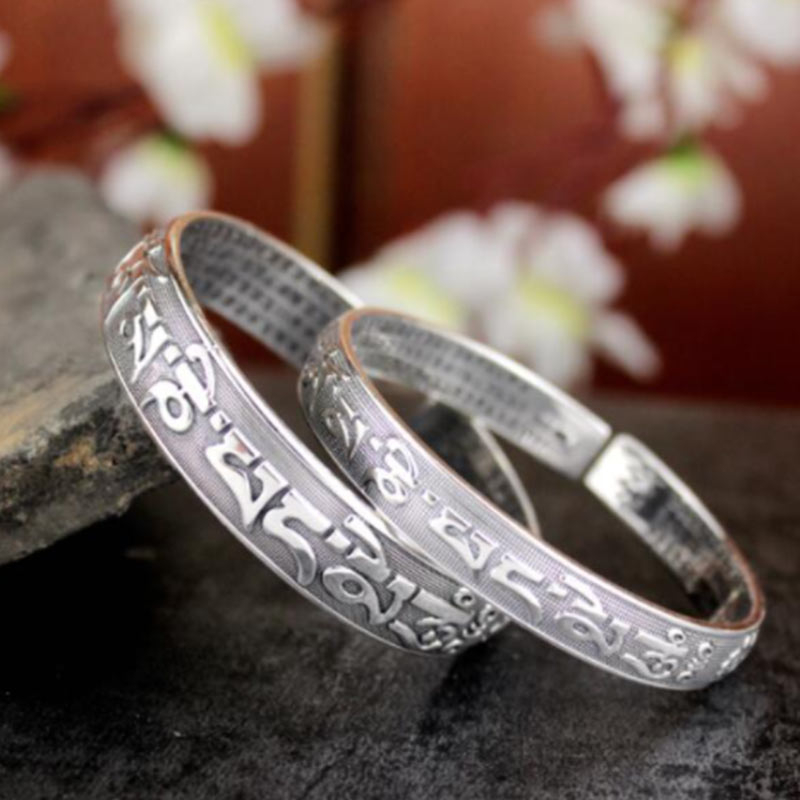 Two Tibetan Om Mani Padme Hum silver prayer bangles overlapping on dark stone surface with raised mantra exterior and micro-engraved interior.