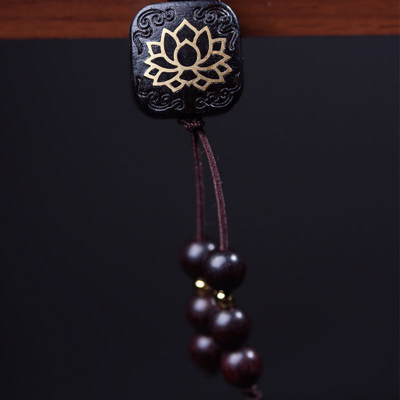 Close-up of square dark wood guru bead with gold lotus flower inlay and hanging tassel with graduated wooden beads on black background