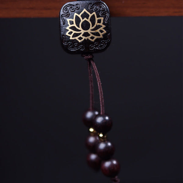 Close-up of square dark wood guru bead with gold lotus flower inlay and hanging tassel with graduated wooden beads on black background