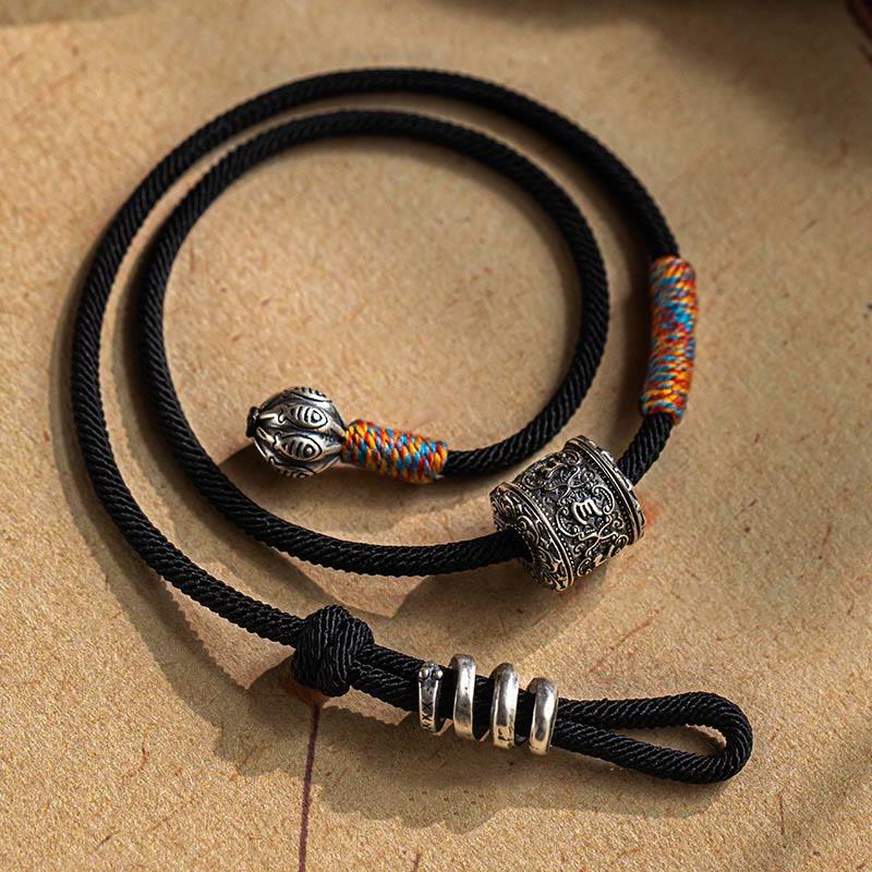 Tibetan Om Mani Padme Hum Wrap Bracelet – Black cord with silver mantra bead and colorful woven accents, coiled on beige textured surface