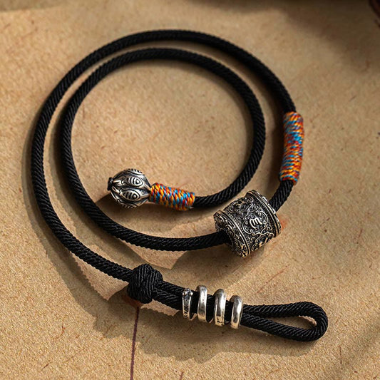 Tibetan Om Mani Padme Hum Wrap Bracelet – Black cord with silver mantra bead and colorful woven accents, coiled on beige textured surface