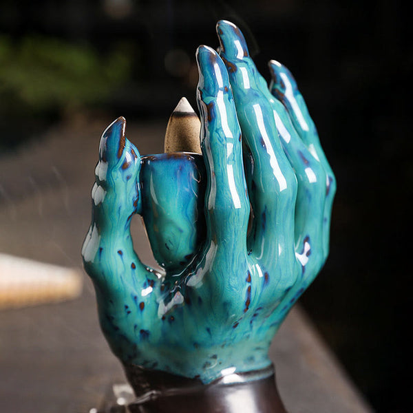 Ceramic Ghost Hand Skull Incense Burner – Turquoise claw hand holding incense cone, wisps of smoke, garden background