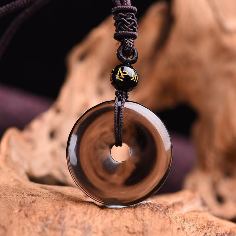 Translucent smoky brown obsidian ping'an kou pendant necklace with black gold-lettered mantra bead on braided cord resting on natural driftwood