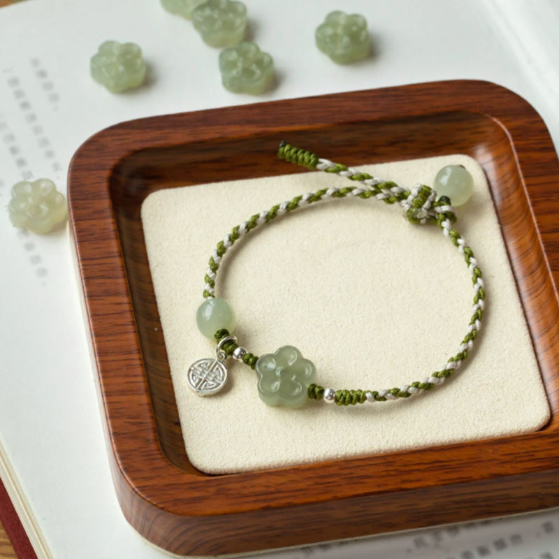 Plum Blossom Jade Flower Braided Bracelet – Green white braided cord with celadon jade flower bead and silver spacer beads on cream fabric in wooden tray, overhead angle