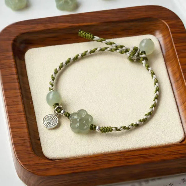Plum Blossom Jade Flower Braided Bracelet – Pale green jade flower bracelet with olive cord and silver charm on cream linen in square wooden tray, top-down view