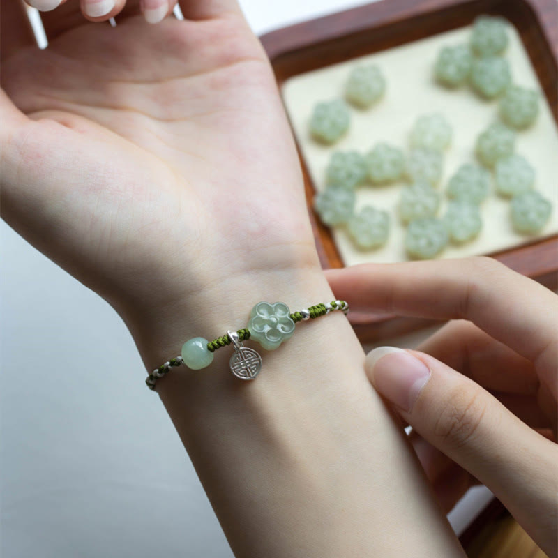 Plum Blossom Jade Flower Braided Bracelet – Green jade flower bracelet worn on wrist with scattered jade flowers on cream tray in background, close-up side view