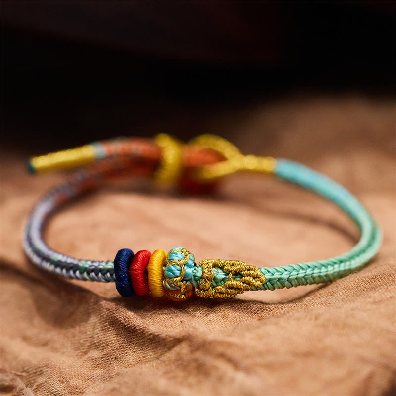 Five-Color Eight Thread Peace Knot Bracelet – Gradient turquoise gray multicolor woven cord with gold yellow red blue decorative knots, tan fabric low side angle